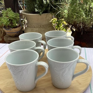 Puede incluir: Seis tazas de cerámica blanca con un diseño floral en relieve dispuestas sobre una tabla redonda de madera. Las tazas tienen asas curvas. El fondo presenta plantas en macetas.
