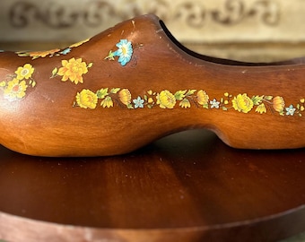 May include: A wooden clog with a floral design painted on the side. The clog is brown and has a yellow, blue, and white floral pattern. The clog is sitting on a wooden base.