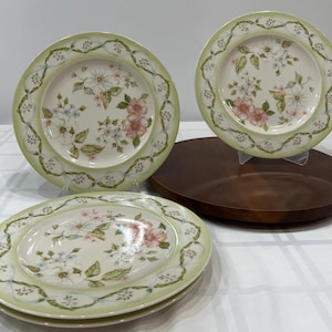 Puede incluir: Un juego de platos decorados con flores. Los platos presentan un borde verde claro con un diseño floral y un arreglo floral central en rosa, blanco y azul. El juego incluye platos redondos y ovalados, perfectos para una mesa.