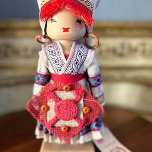 May include: A wooden doll wearing a traditional folk costume with a white head scarf, red and white trim, and a red and white embroidered dress. The doll is holding a red and white fabric square with orange buttons.