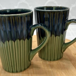May include: Two ceramic mugs with a ribbed design. The mugs are green at the base, transitioning to a dark blue-green at the top. Each mug has a curved handle and sits on a wooden surface.