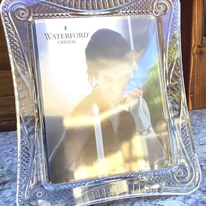 Puede incluir: Un marco de fotos de cristal transparente Waterford con un centro rectangular y bordes moldeados decorativos que presentan volutas y patrones de corte de diamante. El marco contiene un inserto promocional con el texto Waterford Crystal.