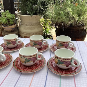 Puede incluir: Un juego de seis tazas y platillos de cerámica blanca. Las tazas y los platillos tienen un patrón de mosaico rojo. Las tazas presentan un diseño de uvas y otras frutas. Los platillos son redondos.