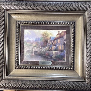 Puede incluir: Una impresión enmarcada de una pintura de Thomas Kinkade titulada "Painter of Light". La pintura representa una pintoresca escena de pueblo con un puente sobre un río, casas con ventanas iluminadas y exuberante vegetación. El marco es de madera marrón oscuro con un acabado dorado.