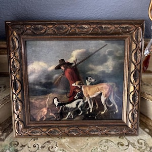 Puede incluir: Una pintura enmarcada que representa a un hombre con un abrigo rojo y un sombrero de ala ancha, sosteniendo un rifle y caminando con tres perros. La pintura está ambientada en un paisaje con un cielo nublado.