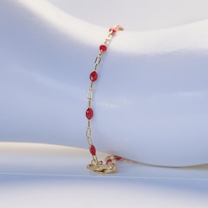 May include: A delicate gold chain bracelet featuring small, evenly spaced red beads. The bracelet is constructed with thin, rectangular gold links alternating with vibrant red, oval-shaped beads, finished with a small gold lobster claw clasp.