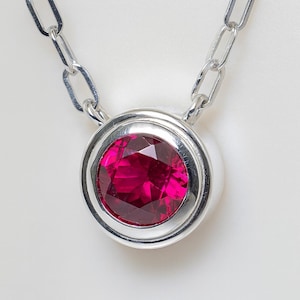 May include: A silver necklace featuring a large, round, faceted ruby pendant. The pendant is set in a bezel setting and hangs from a silver chain with elongated links. The ruby is a vibrant shade of red.