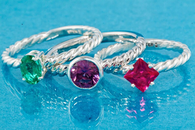 K&ouml;nnte beinhalten: Drei Silberringe mit verschiedenen Edelsteinen. Der Ring links hat einen gr&uuml;nen Smaragd, der mittlere Ring einen violetten Amethyst und der Ring rechts einen roten Rubin.