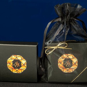K&ouml;nnte beinhalten: Zwei schwarze Schachteln mit goldenen Verzierungen und einem geometrischen Design mit den Buchstaben "JG" darin. Die Schachteln befinden sich auf einem blauen Hintergrund und eine befindet sich in einem schwarzen Netzbeutel mit einem goldenen Band.