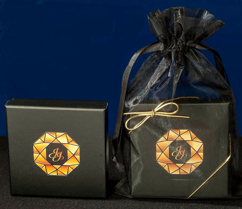 K&ouml;nnte beinhalten: Zwei schwarze Schachteln mit goldenen Verzierungen und einem geometrischen Design mit den Buchstaben "JG" darin. Die Schachteln befinden sich auf einem blauen Hintergrund und eine befindet sich in einem schwarzen Netzbeutel mit einem goldenen Band.