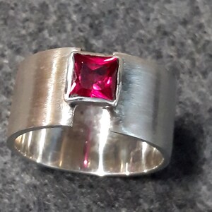 May include: A silver ring with a square, bright pink gemstone set in the center. The ring is wide and has a unique, asymmetrical design.