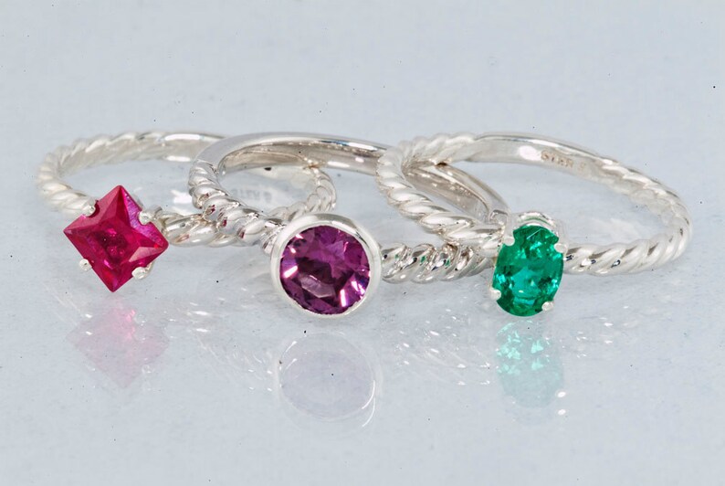K&ouml;nnte beinhalten: Drei Silberringe mit verschiedenen Edelsteinen. Der Ring links hat einen quadratischen roten Edelstein, der mittlere Ring einen runden violetten Edelstein und der Ring rechts einen ovalen gr&uuml;nen Edelstein.