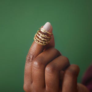 May include: A set of five gold rings stacked on a finger. The top ring has the word "mama" engraved on it. The other rings feature small hearts and gemstones.