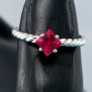 K&ouml;nnte beinhalten: Ein silberner Ring mit einem gedrehten Band und einem quadratischen, leuchtend roten Edelstein.