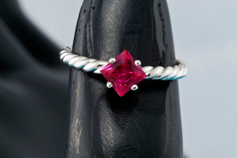 K&ouml;nnte beinhalten: Ein silberner Ring mit einem gedrehten Band und einem quadratischen, leuchtend roten Edelstein.