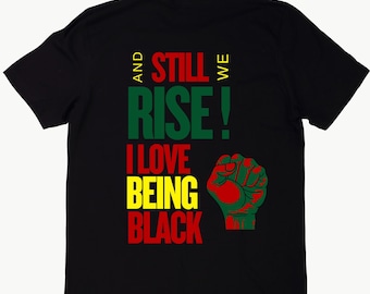 Op de afbeelding: Zwart T-shirt met de tekst "And Still We Rise! I Love Being Black" in rode, groene en gele letters. Een gebalde vuist is afgebeeld in rood, groen en geel.