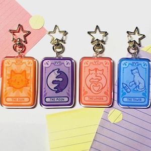 Puede incluir: Cuatro llaveros acrílicos con diseños de cartas del tarot e ilustraciones de gatos. Las cartas son de color naranja, morado, rojo-naranja y azul, cada una con un cierre dorado en forma de estrella. Las cartas están etiquetadas como "The Sun", "The Moon", "The Lovers" y "The Tower".