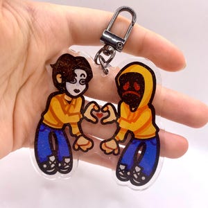 May include: A clear acrylic keyring featuring two cartoon characters wearing yellow hoodies and blue jeans. The characters are positioned facing each other, forming a heart shape with their hands, with a small red heart in the centre.