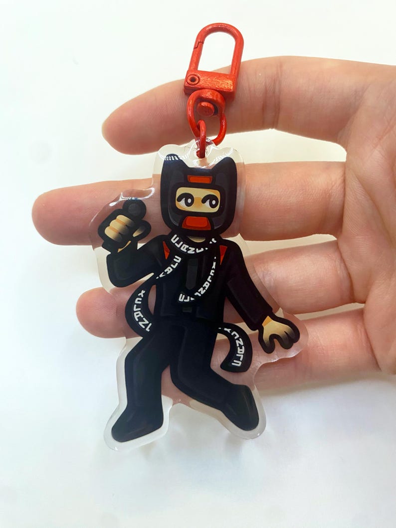以下が含まれることがあります： 赤いマスクと黒いジャケットを着た黒と赤の漫画のキャラクターのキーホルダー。キャラクターは「FREAK」と書かれた黒い尻尾を持っています。キーホルダーは赤い金属製の留め具が付いています。
