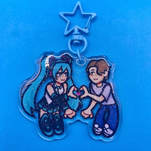 May include: A blue acrylic keyring with a cartoon illustration of two figures making a heart shape with their hands. One figure has long teal hair and the other has brown hair. The keyring has a star-shaped top.