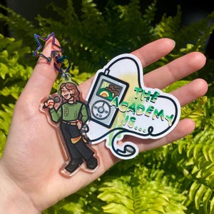 Puede incluir: Un llavero acrílico transparente con un personaje de dibujos animados sosteniendo un micrófono, y una pegatina a juego que dice "The Academy Is..." en verde y blanco. El llavero tiene un cierre en forma de estrella con colores iridiscentes.