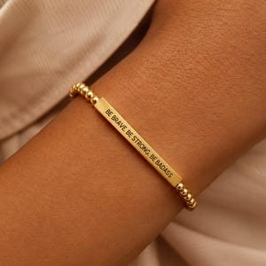 Op de afbeelding: Gouden kralenarmband met een rechthoekige staaf gegraveerd met de woorden "BE BRAVE BE STRONG BE BADASS". De armband wordt om een pols gedragen.