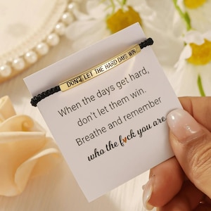 May include: A black braided bracelet with a gold-colored rectangular bar. The bar is engraved with the words "DON'T LET THE HARD DAYS WIN." A white card with inspirational text is behind the bracelet.