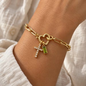 May include: A gold-toned chain bracelet featuring a heart-shaped clasp. Two charms hang from the clasp: a cross encrusted with small clear stones and a rectangular pendant with a single green stone. The bracelet has elongated links.