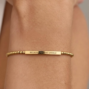 Op de afbeelding: Gouden armband met kleine kralen en een rechthoekige middenstuk. Op het middenstuk staat de tekst "BECAUSE FUCKING SPARKLE". De armband wordt om de arm gedragen.