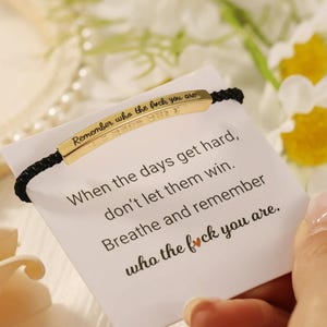 May include: Gold-coloured bracelet with black cord. The bracelet has the engraved text "Remember who the f*ck you are." A card with the text "When the days get hard, don't let them win. Breathe and remember who the f*ck you are."