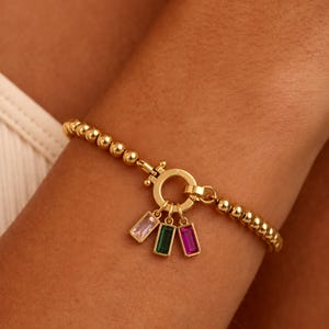 Op de afbeelding: Gouden armband met kralen, een ronde sluiting en drie rechthoekige bedels. De bedels zijn roze, groen en paars. De armband wordt om een arm gedragen.