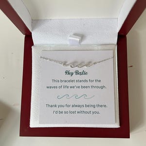 Pode incluir: Um colar prateado em forma de onda apresentado numa caixa bordô com interior branco. Um cartão no interior diz "Hey Bestie" e uma mensagem sobre as ondas da vida. A caixa está aberta, revelando o colar e o cartão.