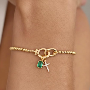 以下が含まれることがあります： ビーズバンドと装飾的な留め金が付いたゴールドトーンのブレスレット。ブレスレットには、長方形のエメラルドグリーンの石と白いアクセントが付いた小さな十字架の2つのチャームが付いています。ブレスレットは手首に着用されています。
