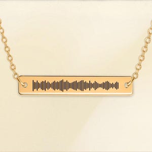 Può includere: Una collana dorata con catena. Il ciondolo rettangolare presenta un motivo a onde sonore nero. La collana è sospesa a una delicata catena, creando un'estetica minimalista ed elegante.