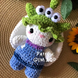 Könnte beinhalten: Ein handgefertigtes weißes Häkelkaninchen mit einem grünen Monsterhut mit drei großen Augen und blauen Latzhosen. Das Kaninchen hat rosa Wangen und schwarze Augen. Der Text auf dem Bild lautet "Patrón: @IM.AQP".