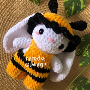 Op de afbeelding: Een gehaakt bij knuffeldier met zwarte en gele strepen, witte vacht en zwart garen haar. Het speelgoed draagt een geel en zwart gestreept overall met de tekst "PATRÓN: @LM.AQP" op de voorkant.