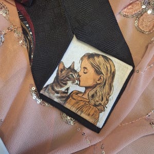 May include: A black tie with a square pattern, featuring a small square patch with an illustration of a girl kissing a tabby cat. The tie has a patterned design on the back and is set against a pink background.