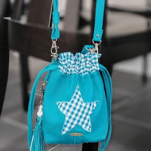 May include: A turquoise drawstring bag featuring a white and blue gingham star. The bag has a white and blue gingham ruffle, a turquoise tassel, and a turquoise shoulder strap. The bag is suspended from a silver chain.