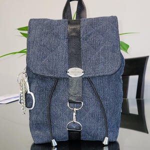 Peut inclure: Un sac à dos en jean avec fermeture à rabat et sangles noires. Le sac à dos comporte une étiquette métallique argentée et un porte-clés avec un charme. Le sac a une poignée noire et des fermetures à cordon.
