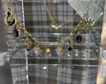 May include: A gold-colored charm necklace featuring various pendants, including a duck, a seahorse, a sunburst, and several gemstone charms. The necklace is displayed on a clear, multi-faceted surface.