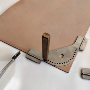 Peut inclure: Un ensemble d'outils de maroquinerie comprenant une pi&egrave;ce de cuir beige, un rainureur de bord en m&eacute;tal, un poin&ccedil;on en m&eacute;tal et un rainureur de couture en m&eacute;tal. Les outils sont dispos&eacute;s sur une surface blanche.