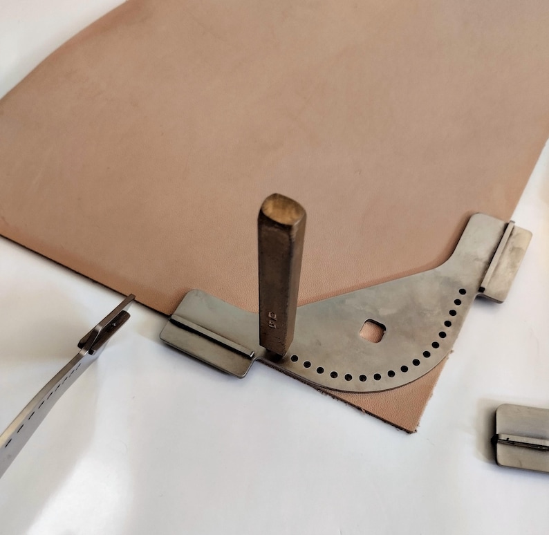 Peut inclure: Un ensemble d'outils de maroquinerie comprenant une pi&egrave;ce de cuir beige, un rainureur de bord en m&eacute;tal, un poin&ccedil;on en m&eacute;tal et un rainureur de couture en m&eacute;tal. Les outils sont dispos&eacute;s sur une surface blanche.