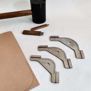 Peut inclure: Des outils de maroquinerie sont dispos&eacute;s sur une surface blanche. Un maillet noir avec un manche en bois est &agrave; c&ocirc;t&eacute; d'outils en m&eacute;tal, notamment des poin&ccedil;ons et des guides de bord incurv&eacute;s. Une pi&egrave;ce de cuir beige avec des trous pr&eacute;-perc&eacute;s est &eacute;galement pr&eacute;sente.
