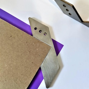 May include: Two silver metal corner brackets with three holes each. The brackets are on a purple and brown surface.