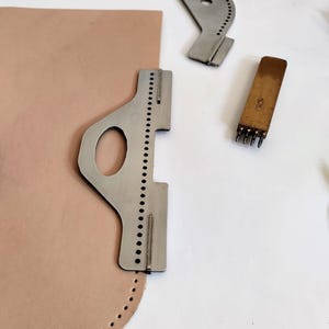 Peut inclure: Des outils de maroquinerie, dont un guide de couture en m&eacute;tal, un poin&ccedil;on &agrave; cuir et un morceau de cuir beige avec des trous pr&eacute;-perc&eacute;s. Les outils sont dispos&eacute;s sur une surface blanche, sugg&eacute;rant un espace de travail pour le travail du cuir.