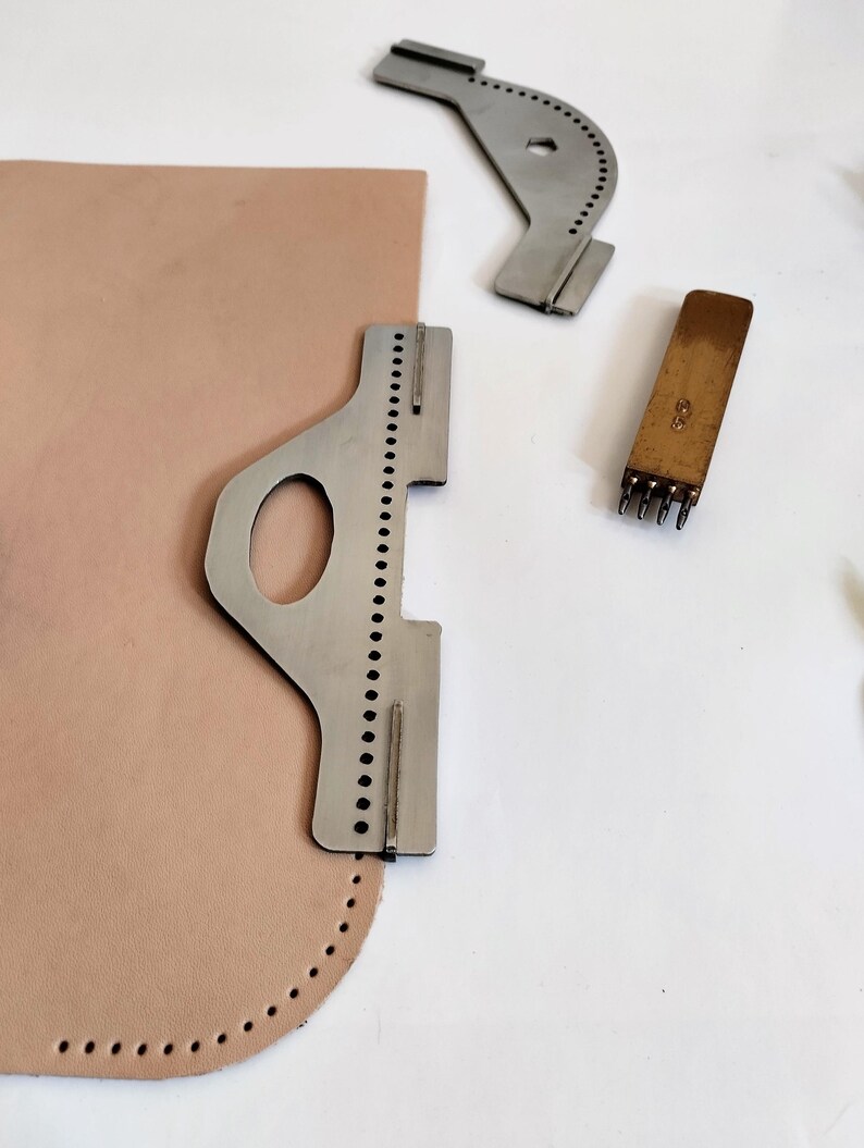 Peut inclure: Des outils de maroquinerie, dont un guide de couture en m&eacute;tal, un poin&ccedil;on &agrave; cuir et un morceau de cuir beige avec des trous pr&eacute;-perc&eacute;s. Les outils sont dispos&eacute;s sur une surface blanche, sugg&eacute;rant un espace de travail pour le travail du cuir.