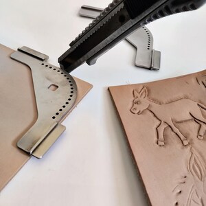 Peut inclure: Gros plan sur des outils et des mat&eacute;riaux de maroquinerie. Un cutter est utilis&eacute; pour couper un morceau de cuir brun clair. Plusieurs gabarits m&eacute;talliques sont visibles, ainsi qu'un morceau de cuir avec un motif d'&acirc;ne en relief.