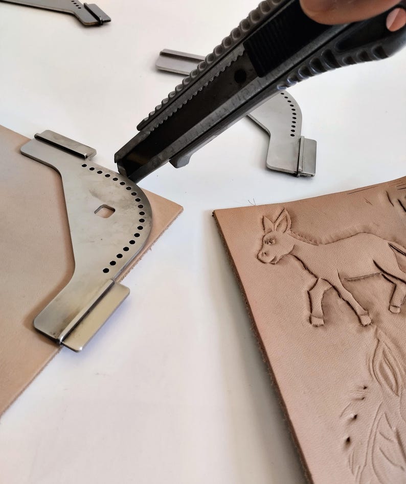 Peut inclure: Gros plan sur des outils et des mat&eacute;riaux de maroquinerie. Un cutter est utilis&eacute; pour couper un morceau de cuir brun clair. Plusieurs gabarits m&eacute;talliques sont visibles, ainsi qu'un morceau de cuir avec un motif d'&acirc;ne en relief.