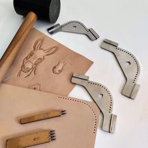 Peut inclure: Ensemble d'outils de travail du cuir, dont un maillet noir, des tampons m&eacute;talliques et des al&egrave;nes &agrave; coudre. Une pi&egrave;ce de cuir beige pr&eacute;sente un motif d'&acirc;ne en relief et des marques de points perfor&eacute;s.