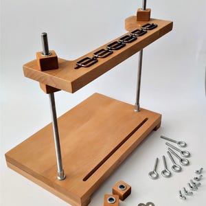 May include: A wooden craft tool with a rectangular base and an angled top piece, connected by metal rods. The top piece has several metal loops. Additional hardware, including eye bolts, washers, and wing nuts, are scattered around the base. The wood has a light brown colour.
